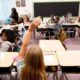Student raises hand in a classroom as a teacher stands at the front.