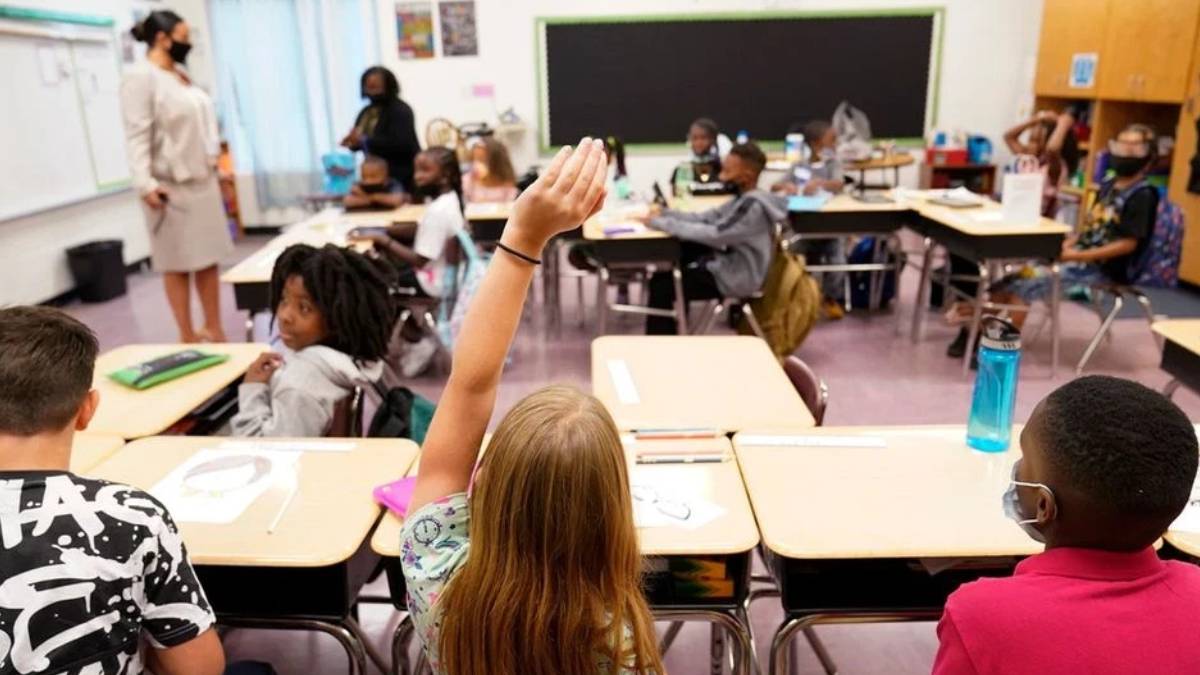 Student raises hand in a classroom as a teacher stands at the front.