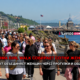 Large group of women walking along a seaside promenade with water and mountains in the background; bold news banner across the center reads Tacoma Girl Walk...
