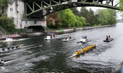 Rowing race on a river beneath a large bridge, crews in yellow uniforms with support boats nearby.