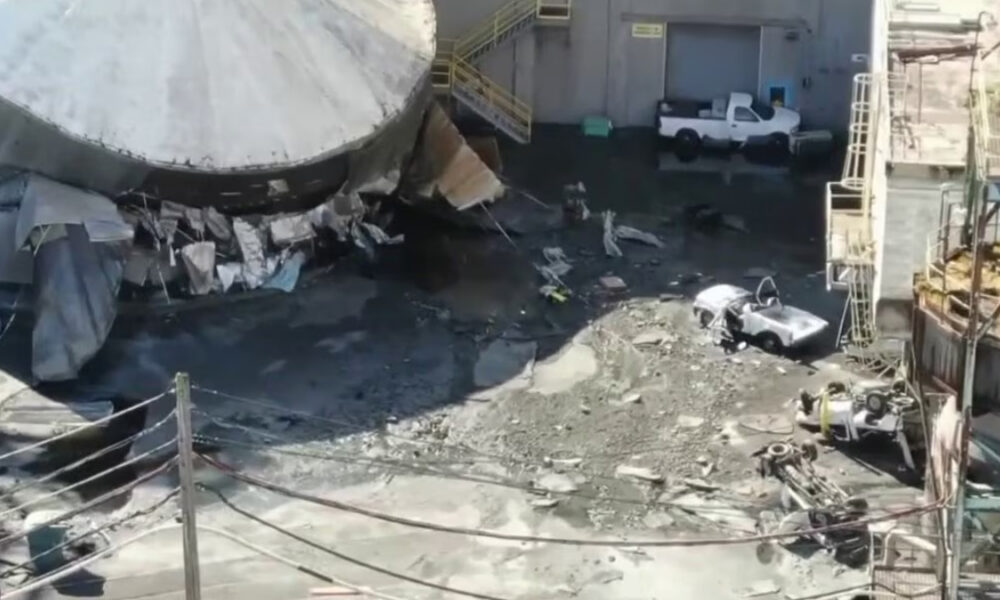 Industrial yard with a partially collapsed large round tank, rubble, and muddy ground; a white pickup and a small silver car near a building.