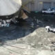 Industrial yard with a partially collapsed large round tank, rubble, and muddy ground; a white pickup and a small silver car near a building.