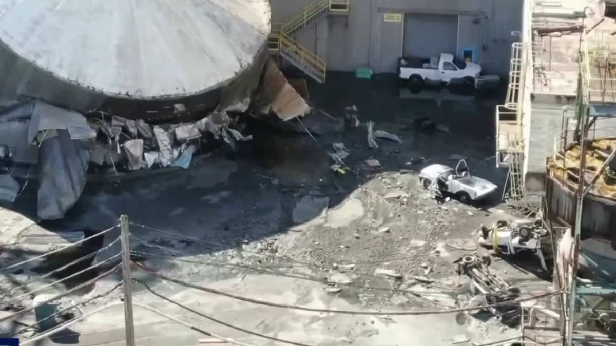 Industrial yard with a partially collapsed large round tank, rubble, and muddy ground; a white pickup and a small silver car near a building.