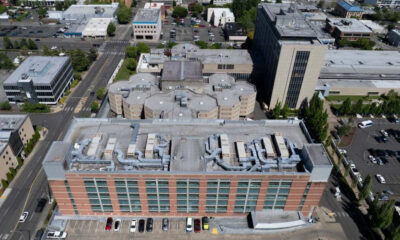 Aerial view of a red-brick office building with large windows and a busy street grid surrounding it, parked cars along the front, and rooftops with HVAC equipment visible.