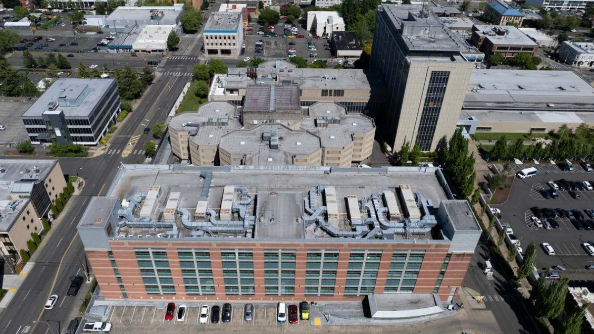 Aerial view of a red-brick office building with large windows and a busy street grid surrounding it, parked cars along the front, and rooftops with HVAC equipment visible.