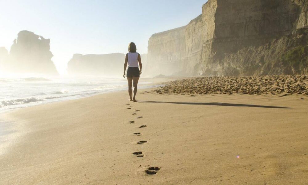 Person walking along a sunlit beach, leaving footprints in the sand with tall cliffs in the background.