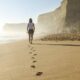 Person walking along a sunlit beach, leaving footprints in the sand with tall cliffs in the background.