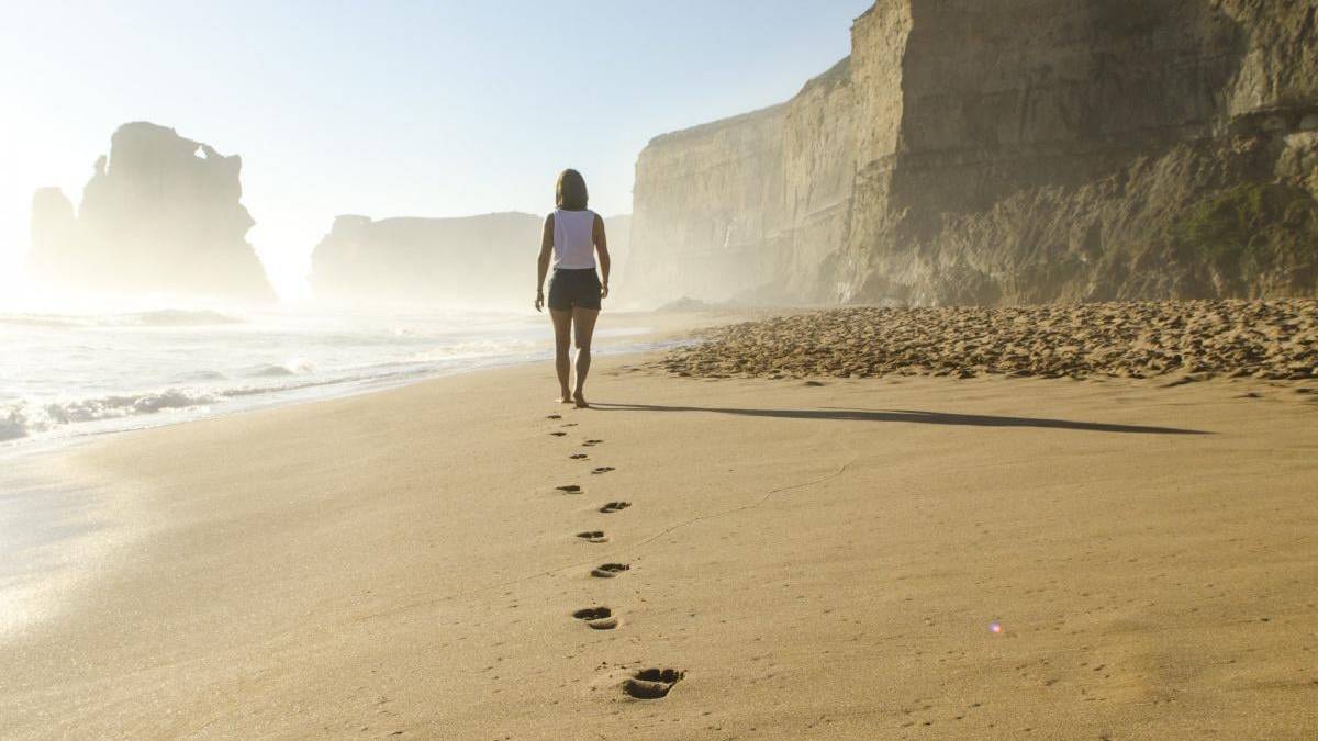 Person walking along a sunlit beach, leaving footprints in the sand with tall cliffs in the background.