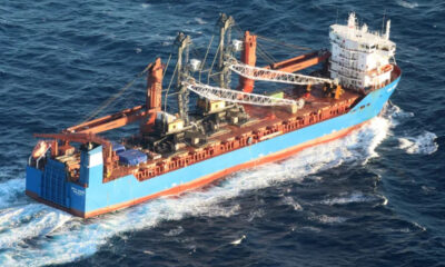 Large blue cargo ship with multiple cranes on deck navigating choppy open water.