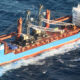 Large blue cargo ship with multiple cranes on deck navigating choppy open water.