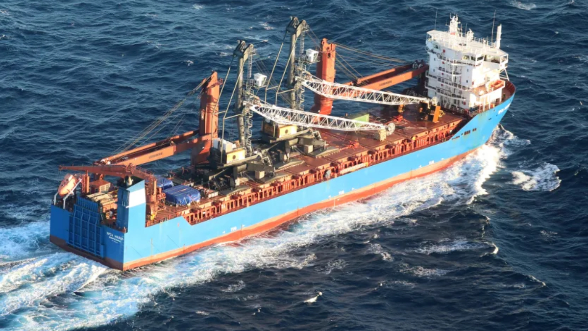 Large blue cargo ship with multiple cranes on deck navigating choppy open water.