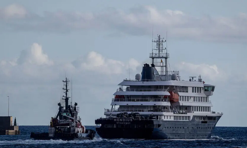 A large multi-deck cruise ship being guided by a tugboat in open water near a breakwater.