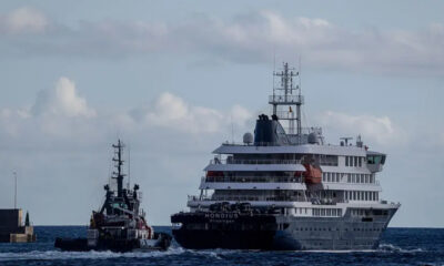 A large multi-deck cruise ship being guided by a tugboat in open water near a breakwater.