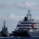 A large multi-deck cruise ship being guided by a tugboat in open water near a breakwater.