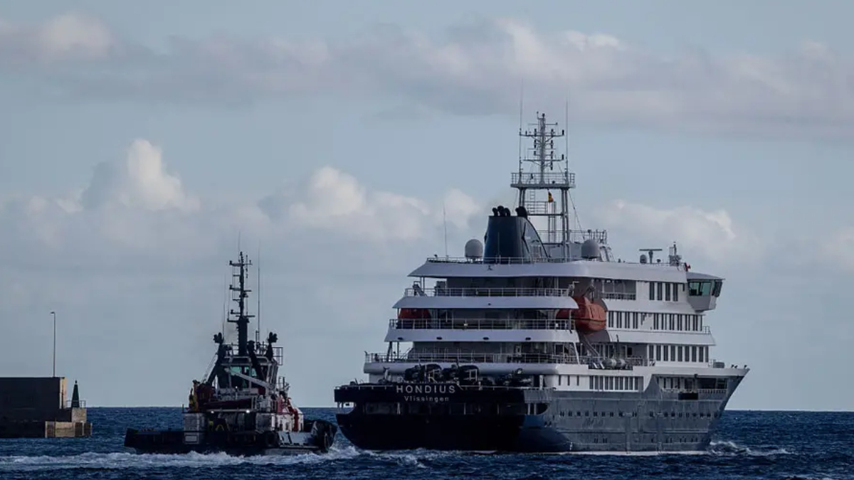A large multi-deck cruise ship being guided by a tugboat in open water near a breakwater.