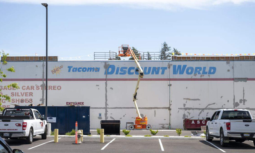 A storefront wall with a large faded sign reading 'Tacoma Discount World' while an orange boom lift works on the roof.