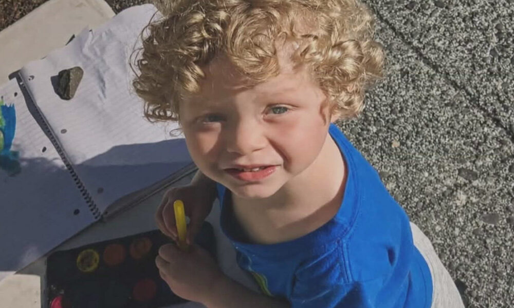 Young boy with curly blond hair in a blue shirt, holding a yellow marker, looking up at the camera outdoors.