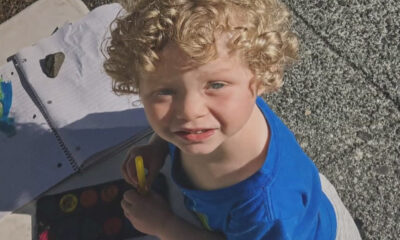 Young boy with curly blond hair in a blue shirt, holding a yellow marker, looking up at the camera outdoors.
