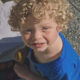 Young boy with curly blond hair in a blue shirt, holding a yellow marker, looking up at the camera outdoors.