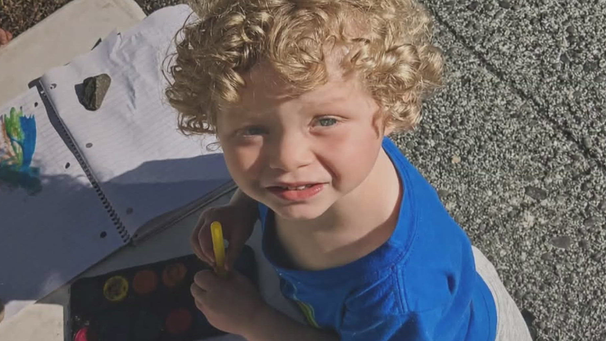 Young boy with curly blond hair in a blue shirt, holding a yellow marker, looking up at the camera outdoors.