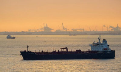 Large dark tanker ship on calm water at sunset with hazy port cranes in the distance on the horizon.