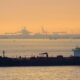 Large dark tanker ship on calm water at sunset with hazy port cranes in the distance on the horizon.