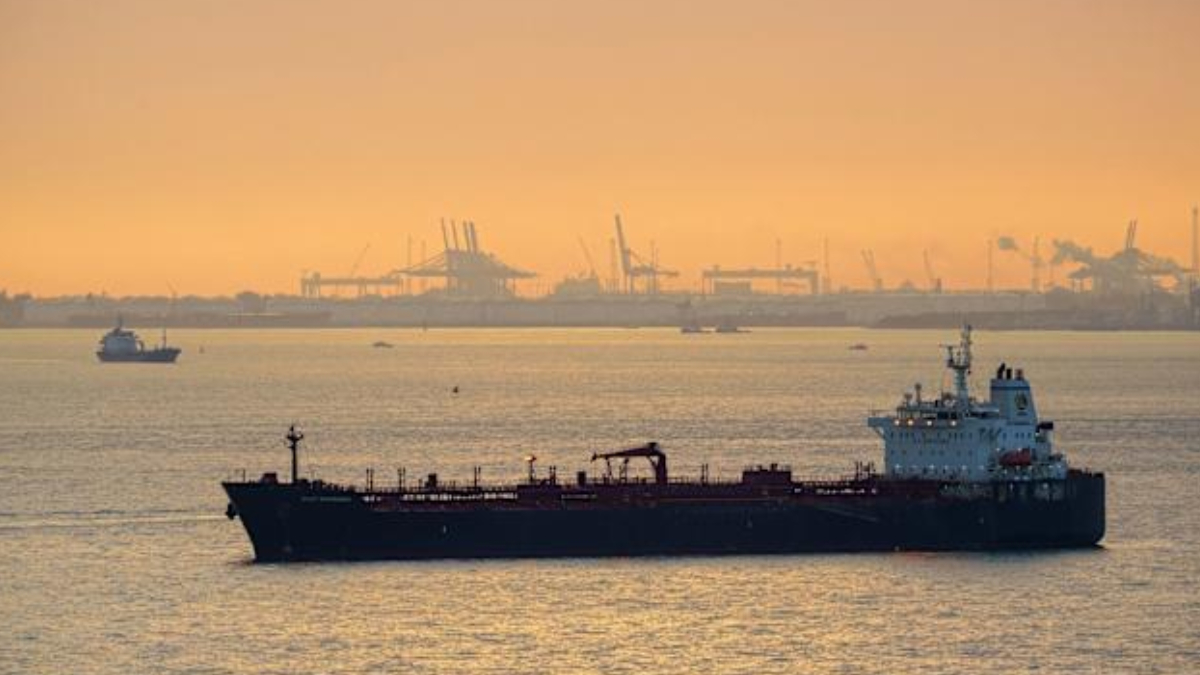 Large dark tanker ship on calm water at sunset with hazy port cranes in the distance on the horizon.
