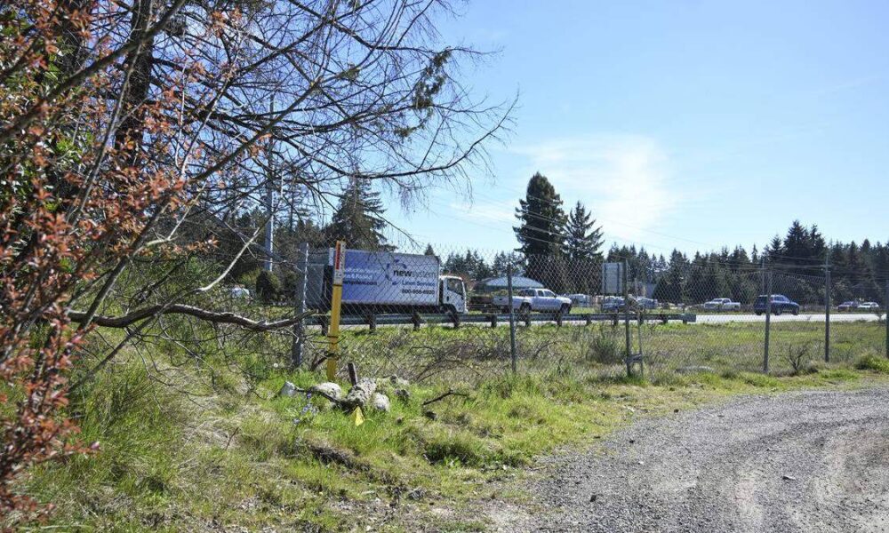 Chain-link fence beside a gravel lot with a line of cars on a highway in the background under a clear blue sky.