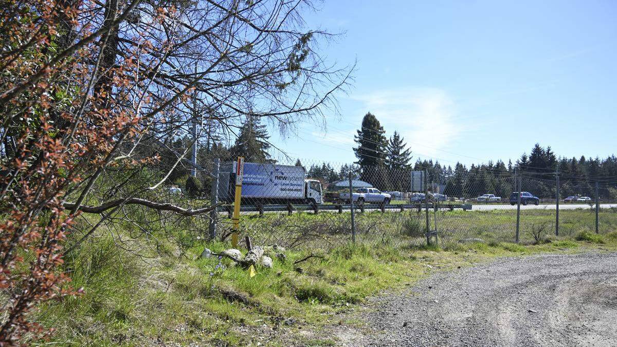 Chain-link fence beside a gravel lot with a line of cars on a highway in the background under a clear blue sky.