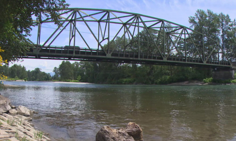 Steel truss bridge spans a calm river with trees along both banks and a rocky shoreline in the foreground.