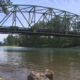 Steel truss bridge spans a calm river with trees along both banks and a rocky shoreline in the foreground.