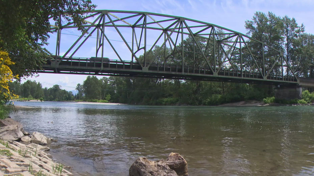 Steel truss bridge spans a calm river with trees along both banks and a rocky shoreline in the foreground.