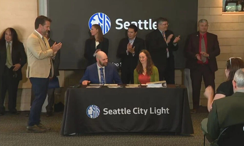 Two people sign documents at a table with a Seattle City Light banner; a group of professionals applaud in the background during a formal ceremony with a Seattle logo on a screen behind them.