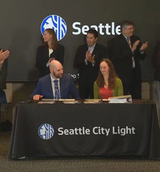 Two people sign documents at a table with a Seattle City Light banner; a group of professionals applaud in the background during a formal ceremony with a Seattle logo on a screen behind them.