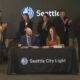 Two people sign documents at a table with a Seattle City Light banner; a group of professionals applaud in the background during a formal ceremony with a Seattle logo on a screen behind them.