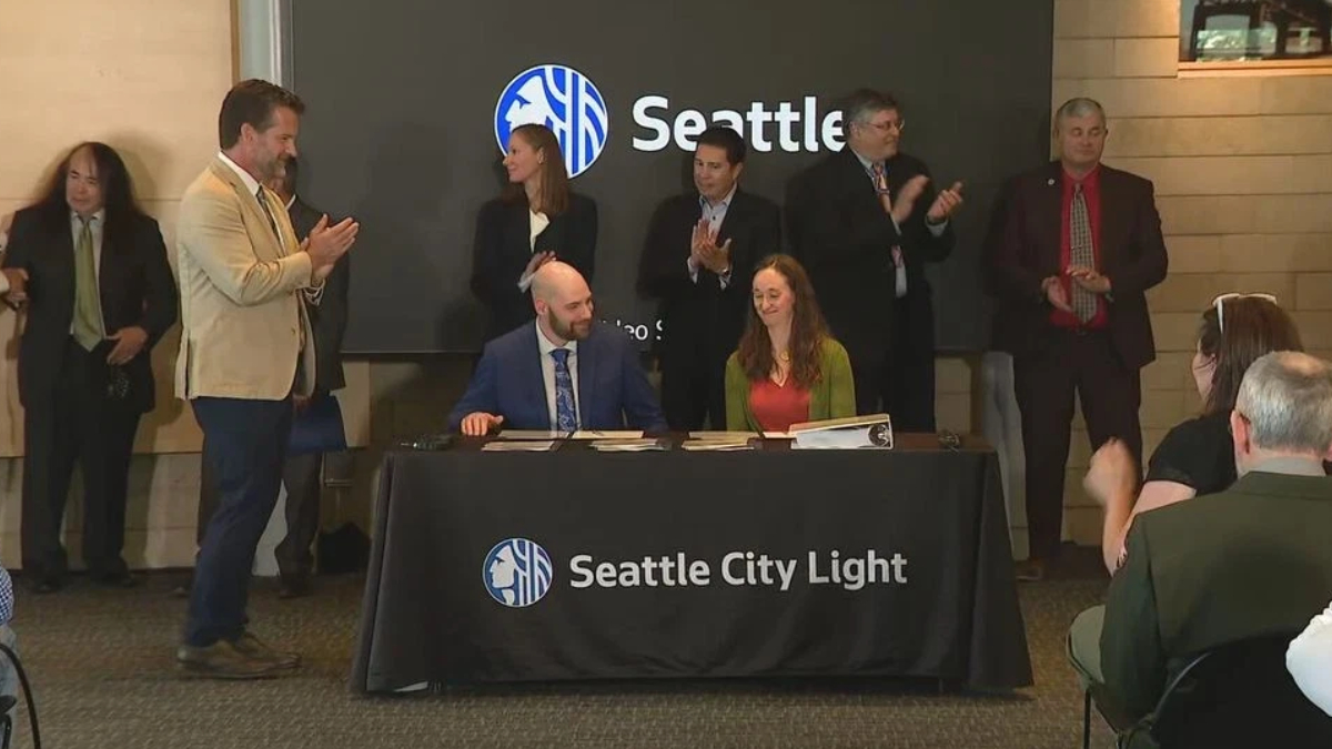 Two people sign documents at a table with a Seattle City Light banner; a group of professionals applaud in the background during a formal ceremony with a Seattle logo on a screen behind them.
