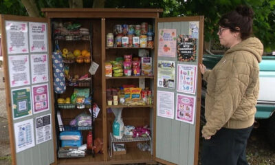 Person in a brown quilted jacket stands beside an open wooden community pantry filled with cans, boxes, and snacks on shelves nearby posters and paperwork on the doors