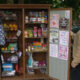 Person in a brown quilted jacket stands beside an open wooden community pantry filled with cans, boxes, and snacks on shelves nearby posters and paperwork on the doors