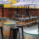 Outdoor beer garden scene with wooden picnic tables under striped purple umbrellas; barrel tables with glass tops and a lone person seated at a table in the background.