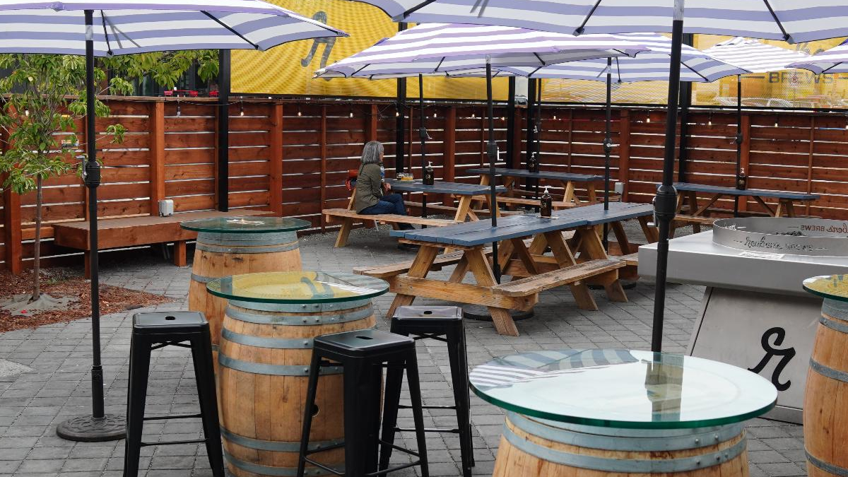 Outdoor beer garden scene with wooden picnic tables under striped purple umbrellas; barrel tables with glass tops and a lone person seated at a table in the background.