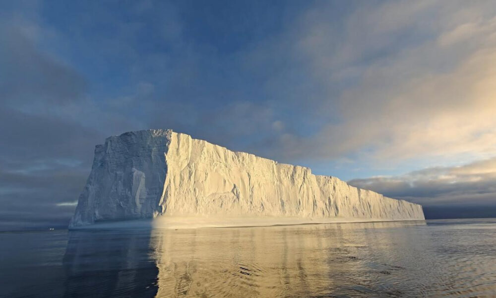 Iceberg rises from a calm sea, its sunlit cliff faces glowing gold as it reflects in the water at sunset.
