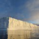Iceberg rises from a calm sea, its sunlit cliff faces glowing gold as it reflects in the water at sunset.