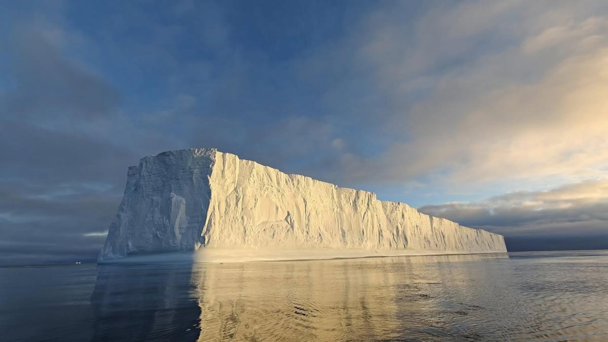 Iceberg rises from a calm sea, its sunlit cliff faces glowing gold as it reflects in the water at sunset.