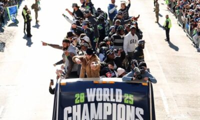 Group of athletes standing and waving from an open-top vehicle during a street parade, banner reads WORLD CHAMPIONS 2025.