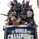 Group of athletes standing and waving from an open-top vehicle during a street parade, banner reads WORLD CHAMPIONS 2025.