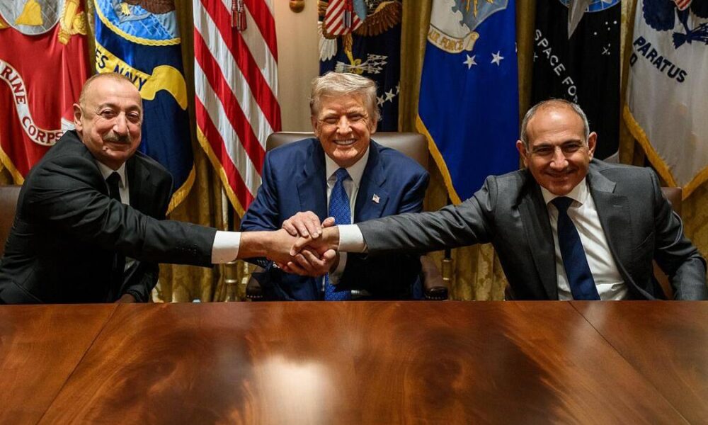 Three men in suits sit at a wood conference table; two on the ends shake hands, smiling, with flags behind them.
