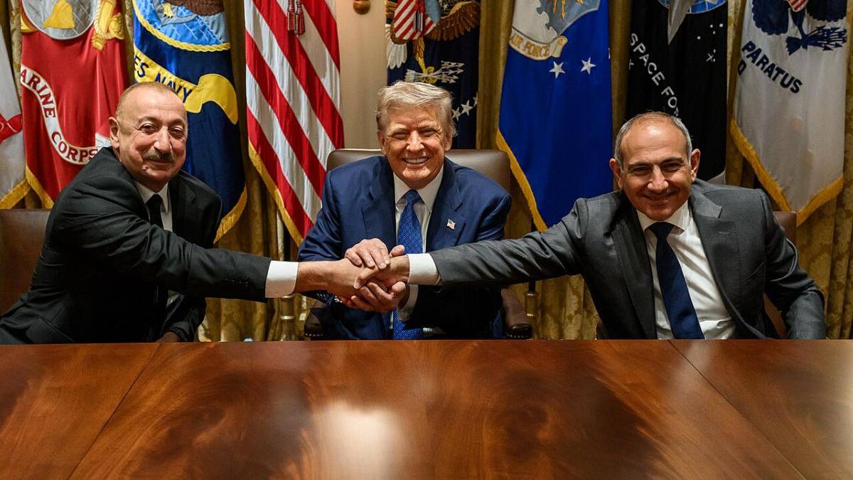 Three men in suits sit at a wood conference table; two on the ends shake hands, smiling, with flags behind them.