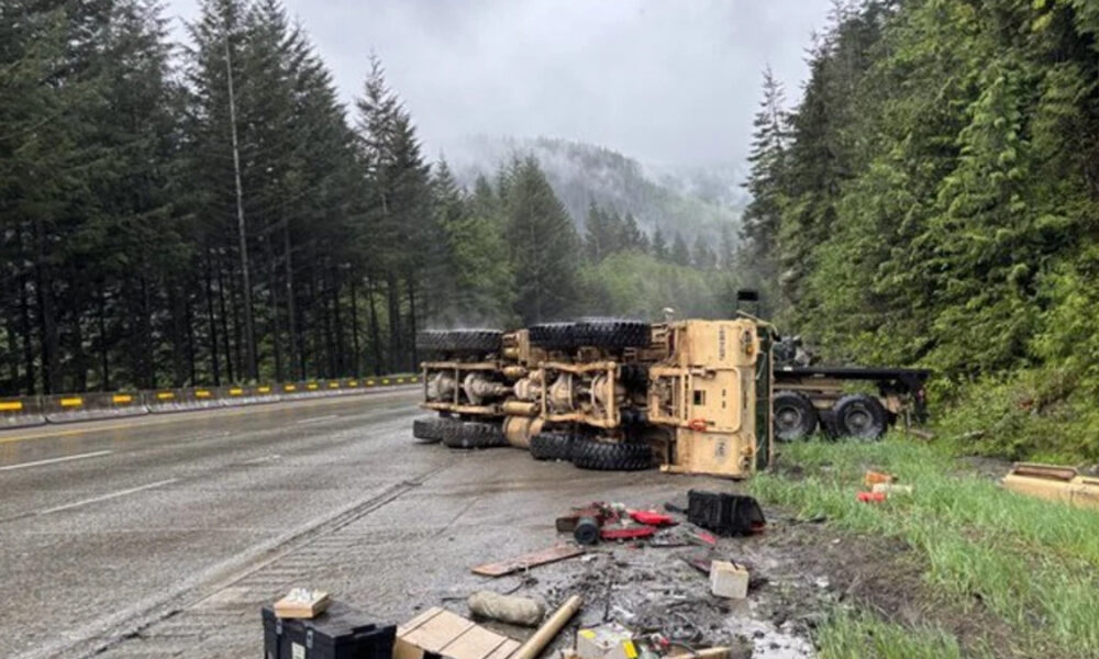 Yellow dump truck lies on its side across a wet forest highway after a crash, with scattered debris along the shoulder.