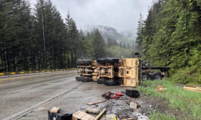 Yellow dump truck lies on its side across a wet forest highway after a crash, with scattered debris along the shoulder.