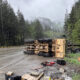 Yellow dump truck lies on its side across a wet forest highway after a crash, with scattered debris along the shoulder.